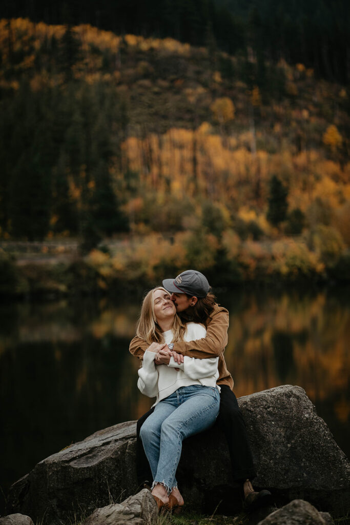 A newly engaged couple holding and kissing each other with the Columbia river and evergreen trees in the background. 