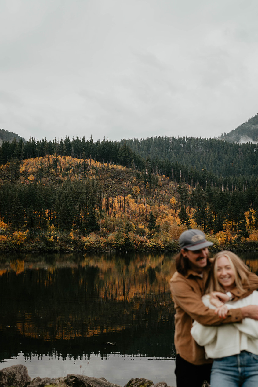 A newly engaged couple holding each other with the Columbia river and evergreen trees in the background. 