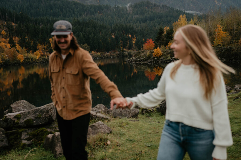 A couple holding hands and looking lovingly at each other with the Columbia river and evergreen trees in the background. 