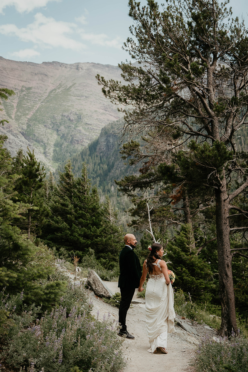 The newlyweds hiking along a trail with pine trees and mountains around them. 