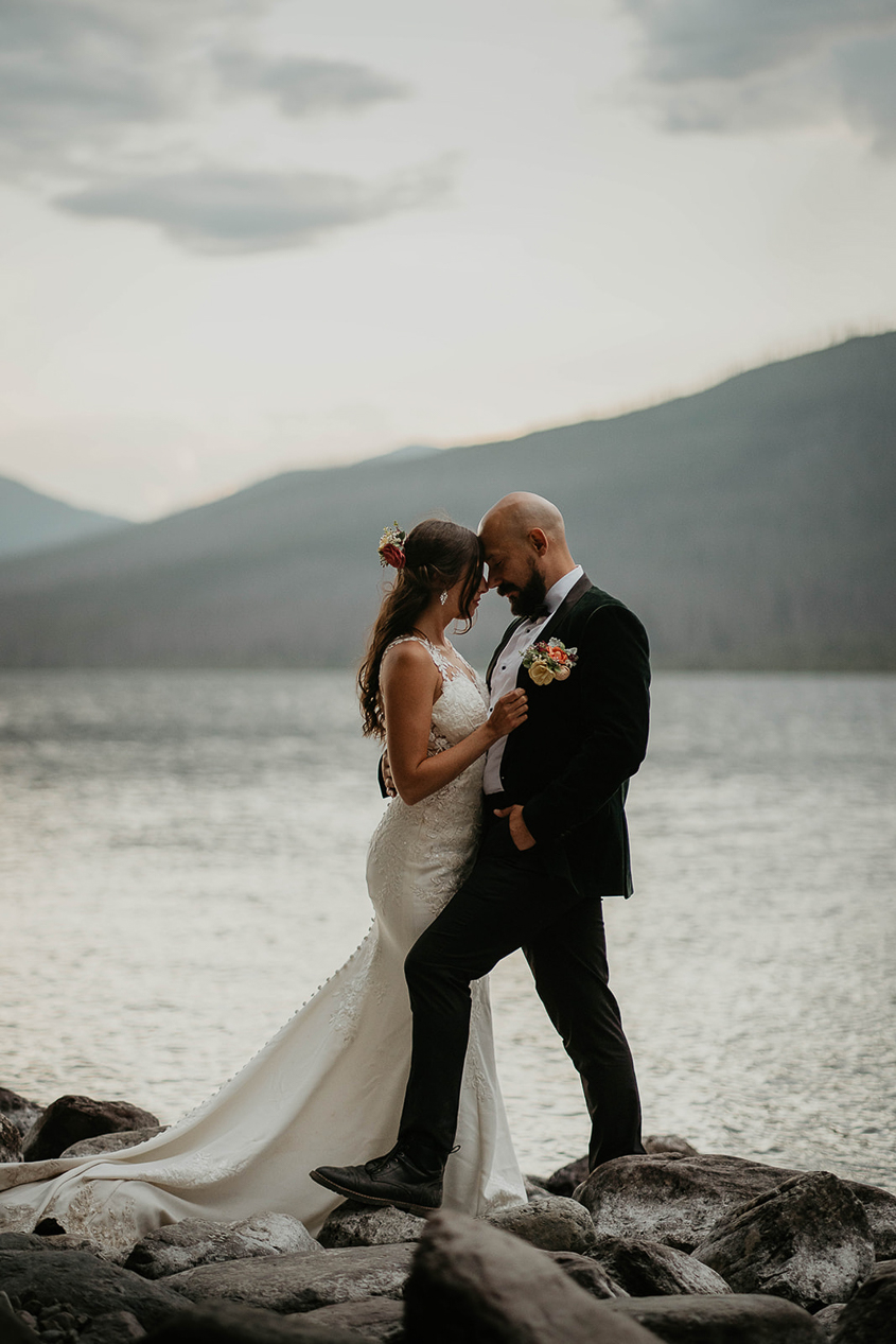 The couple standing close on the lake shore with mountains around them. 