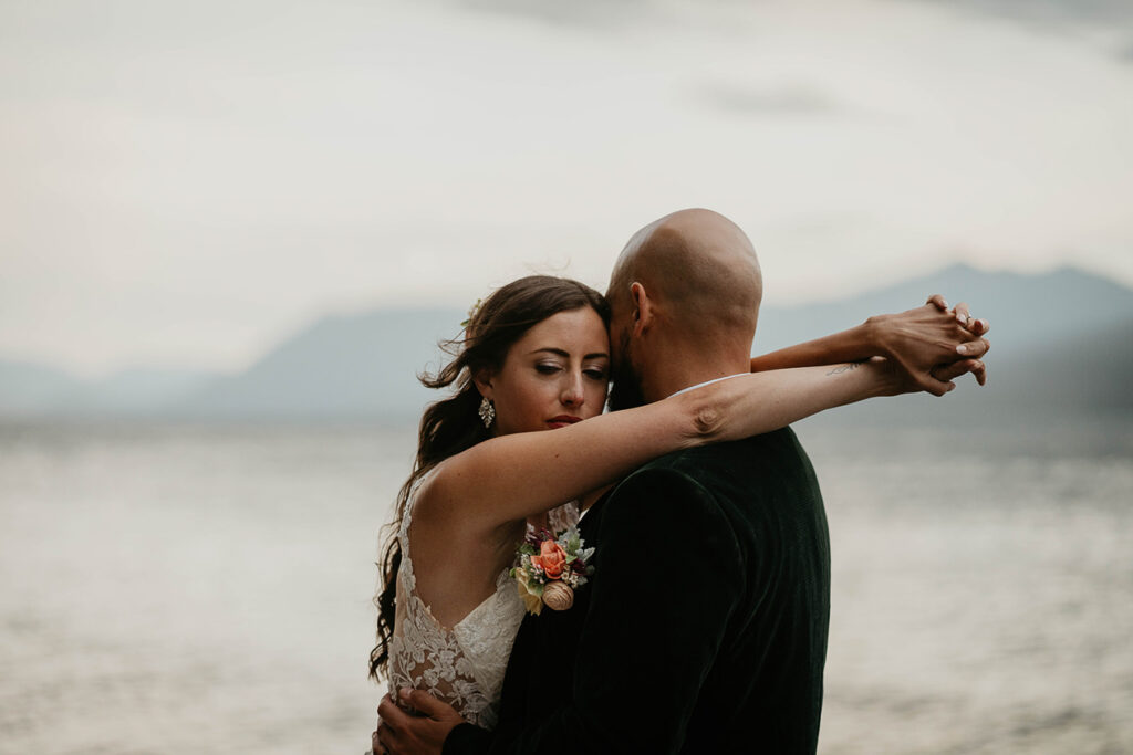 The couple hugging on the lake shore with mountains around them. 