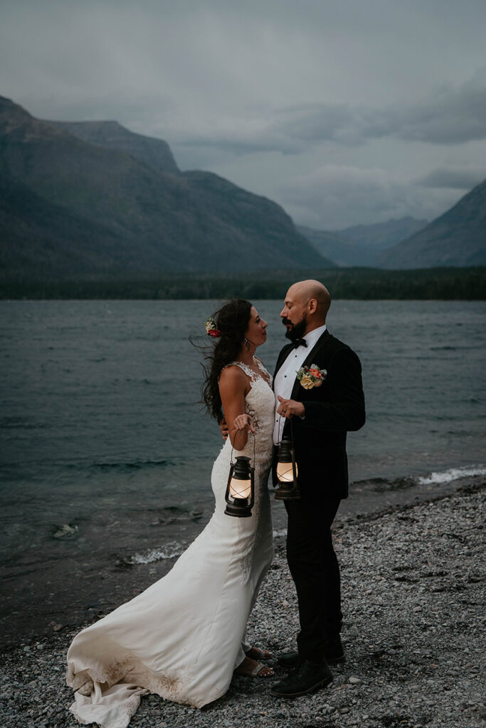 The couple holding lanterns standing on the lake shore with mountains around them and nighttime rolling in. 