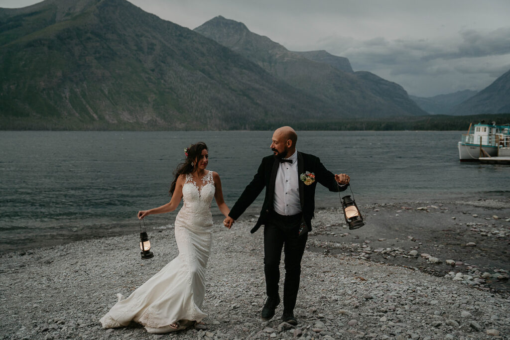 The couple holding lanterns standing on the lake shore with mountains around them and nighttime rolling in. 