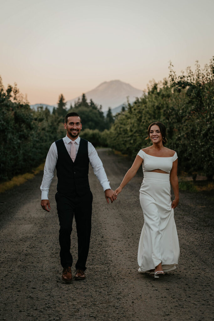 the bride and groom holding hands while walking through a grove of trees at The Orchard Hood River. 