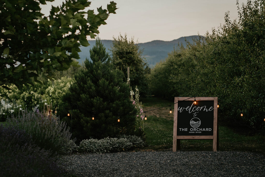 A sign that reads "Welcome to The Orchard Hood River" with lit up string lights, a grove of trees, and flowers. 