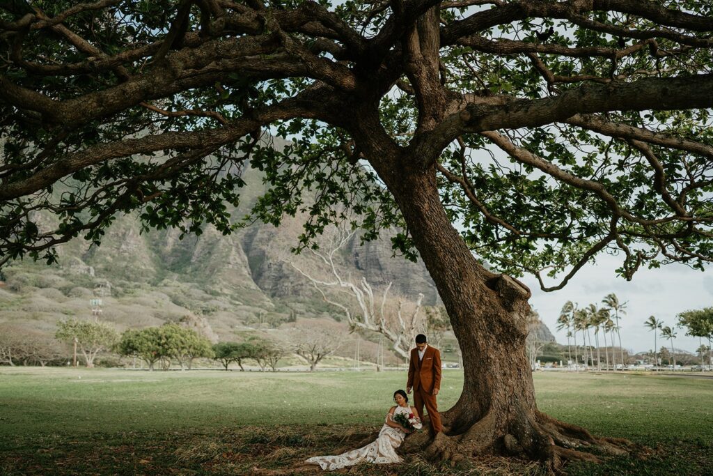 Bride sits on the lawn under a large tree while groom stands nearby during their Hawaii elopement