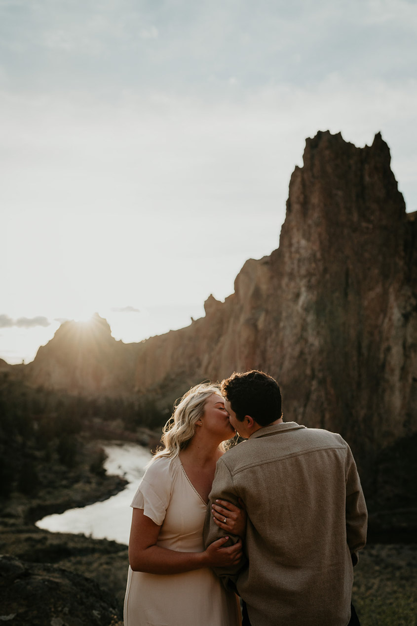 a couple holding each other and kissing with rivers and spires in the background during their Smith Rock engagement session. 