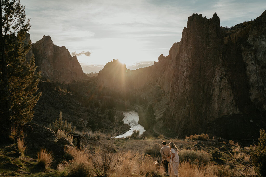 A couple walking through a field with a river and spires in the background during their Smith Rock engagement session. 