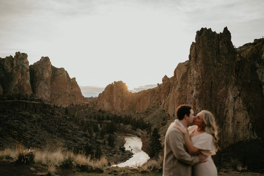 The couple kissing with a river and cliffs in the background during their Smith Rock engagement session. 