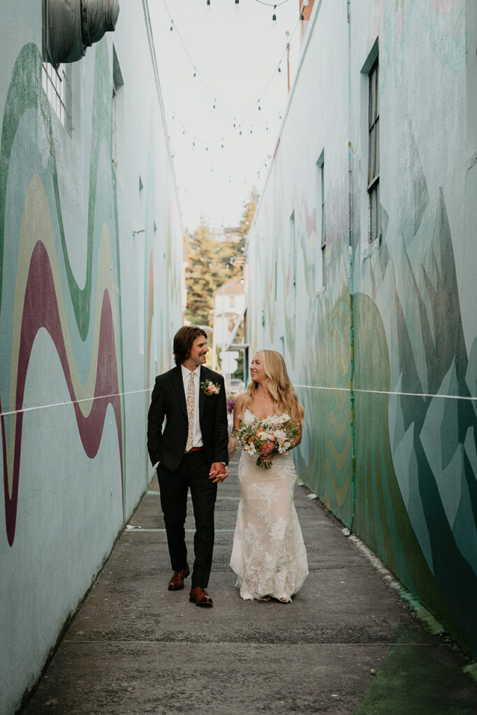 the newlyweds holding hands and walking down an alley way filled with murals. 