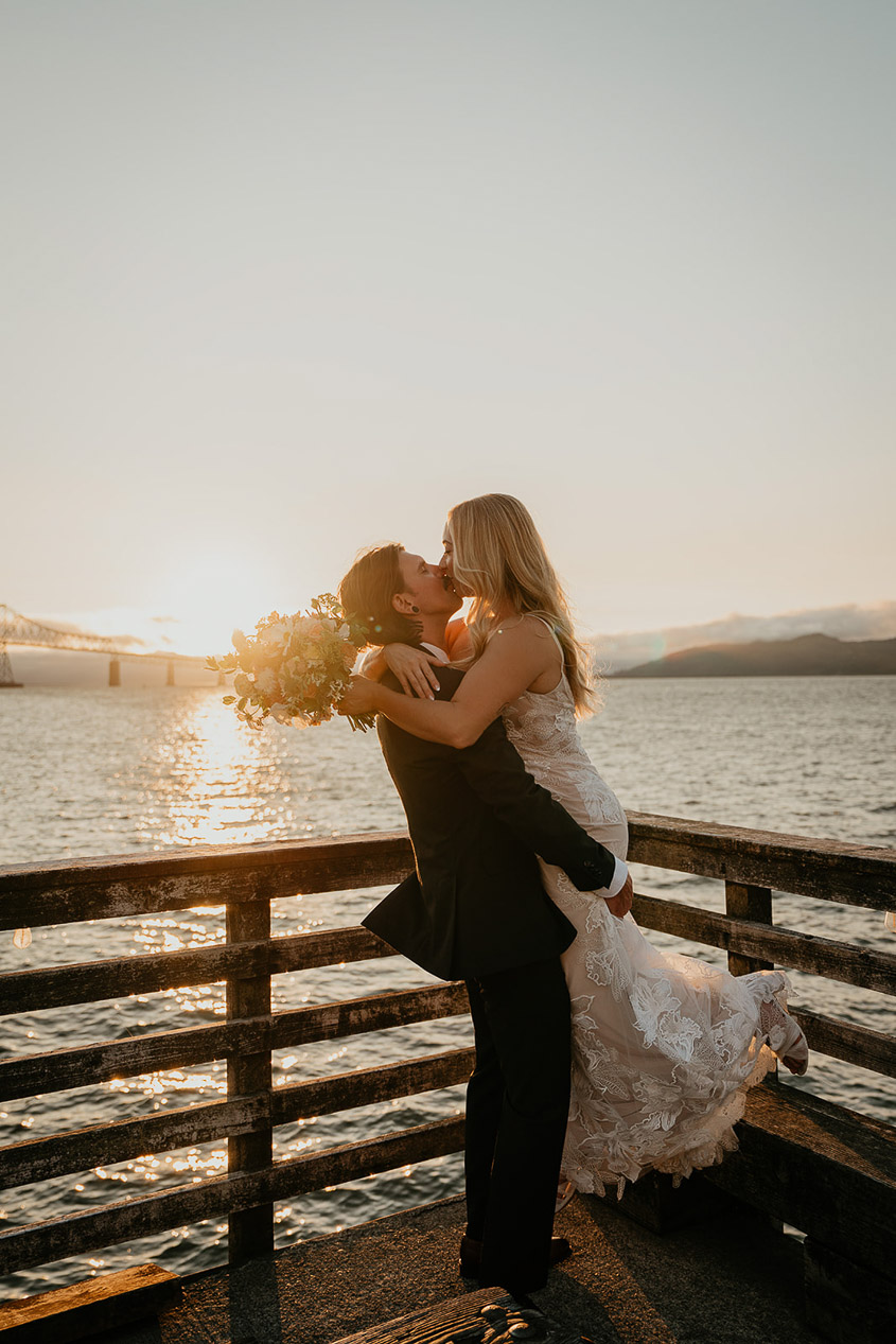 The newlyweds kissing on a dock at sunset.