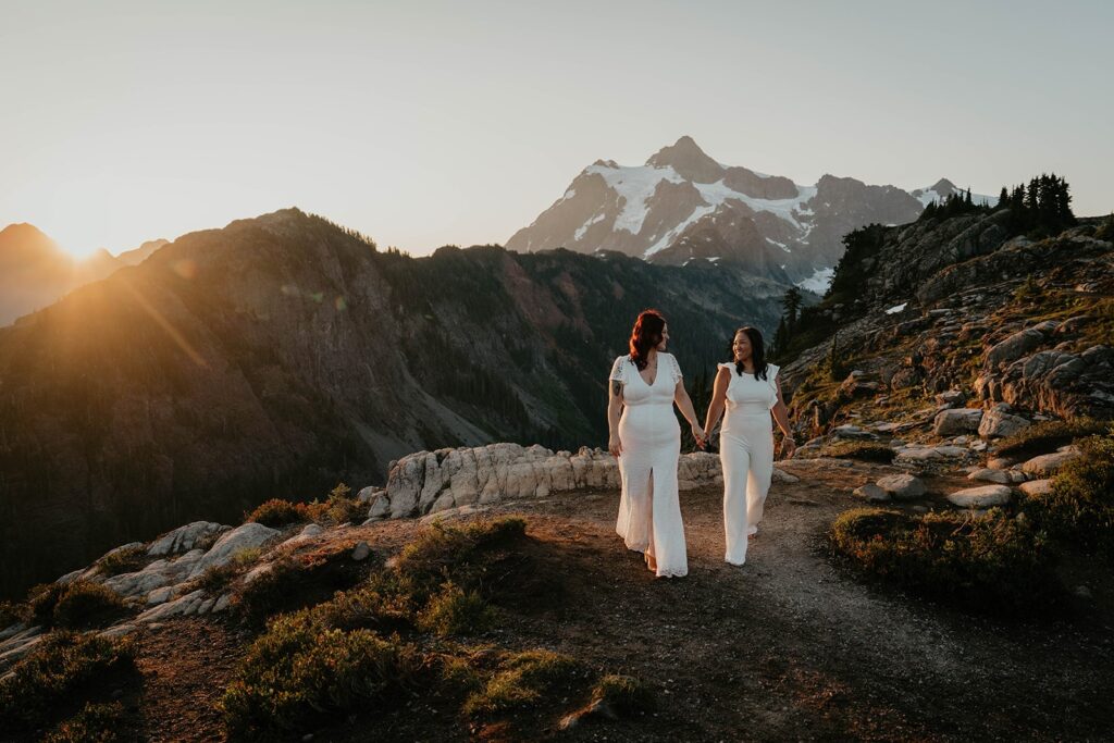 Brides hold hands while walking across a mountain trail during their elopement in the North Cascades