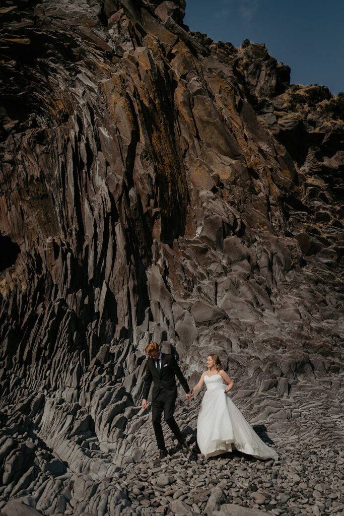 Bride and groom hold hands while walking across a black sand beach while they elope in Iceland 