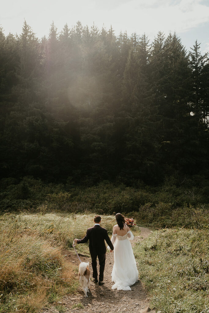 a couple walking their dog on their wedding day though a meadow. 