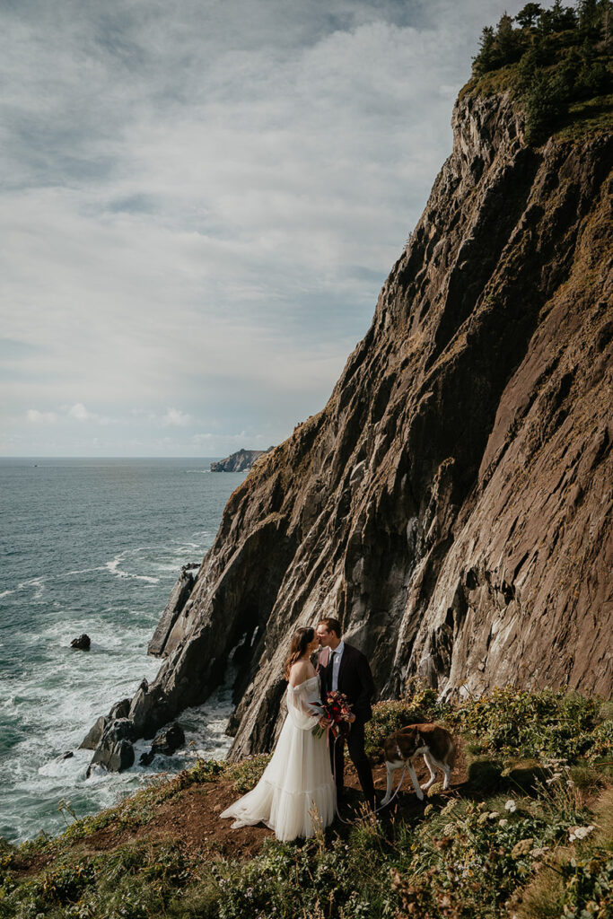 The newlyweds kissing on the Oregon Coast during their elopement while holding their dog. 