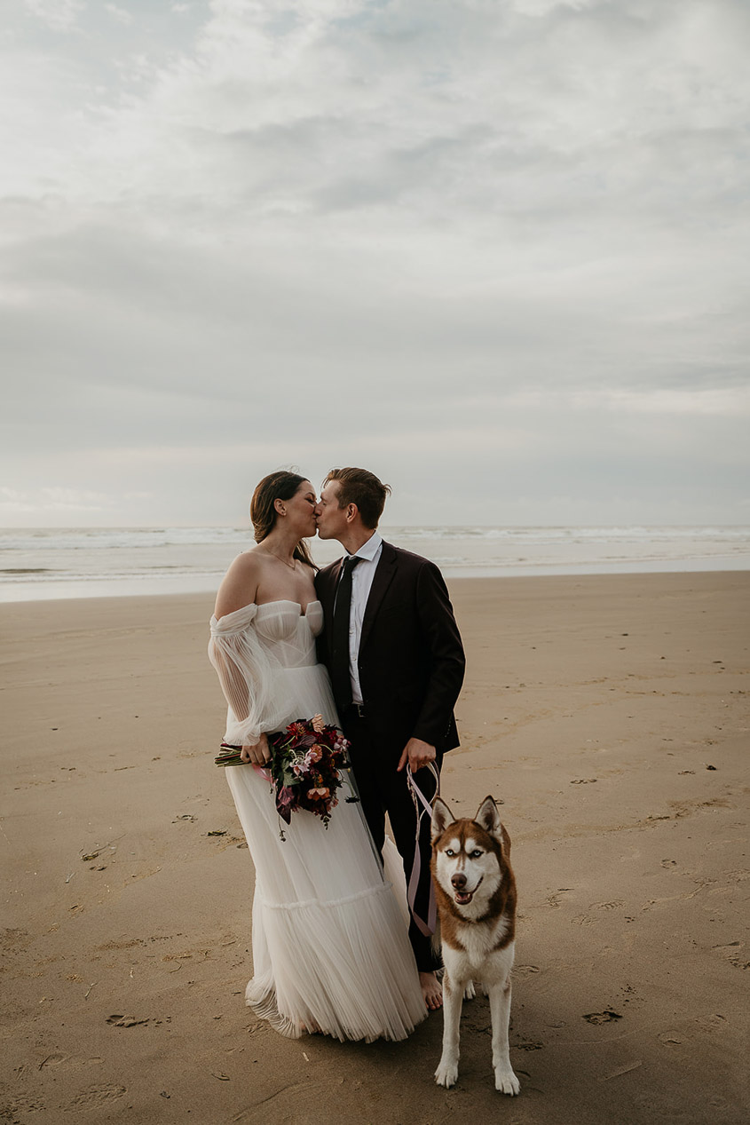 a couple kissing on the Oregon coast while holding their dog on a leash.