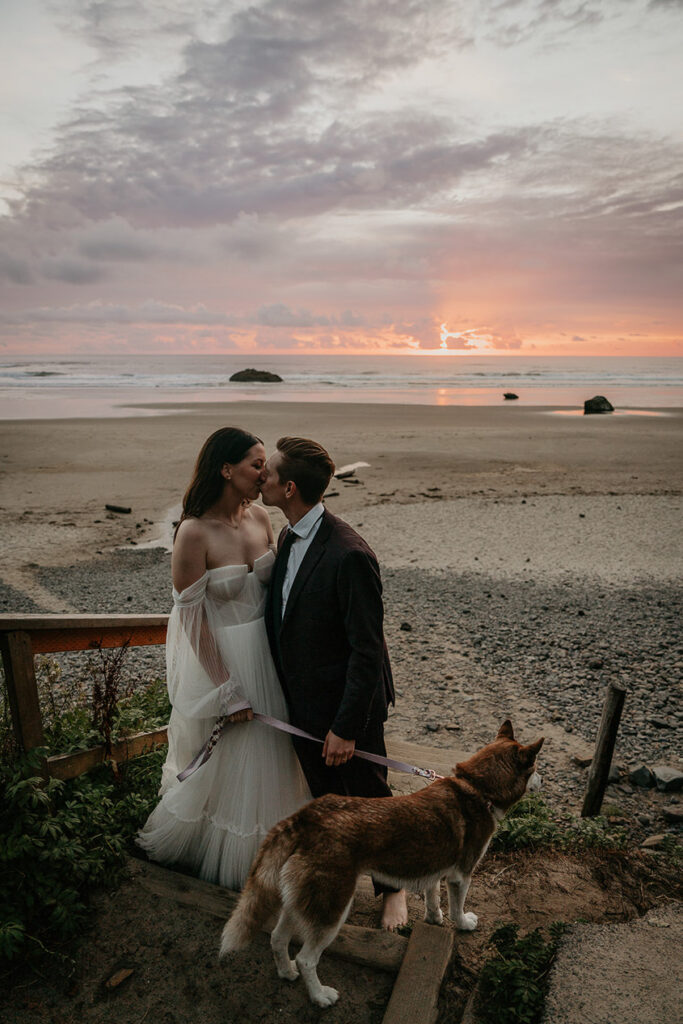 The newlyweds kissing during their Oregon Coast elopement as the sun sets, while holding their dog. 